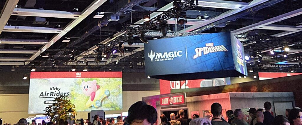 Photograph taken at PAX West 2025, image of the large, digital ceiling-hanging displays for "Kirby Air Riders" and "MTG and Marvel Spider-Man" above the crowds in the area containing the console demo booths.